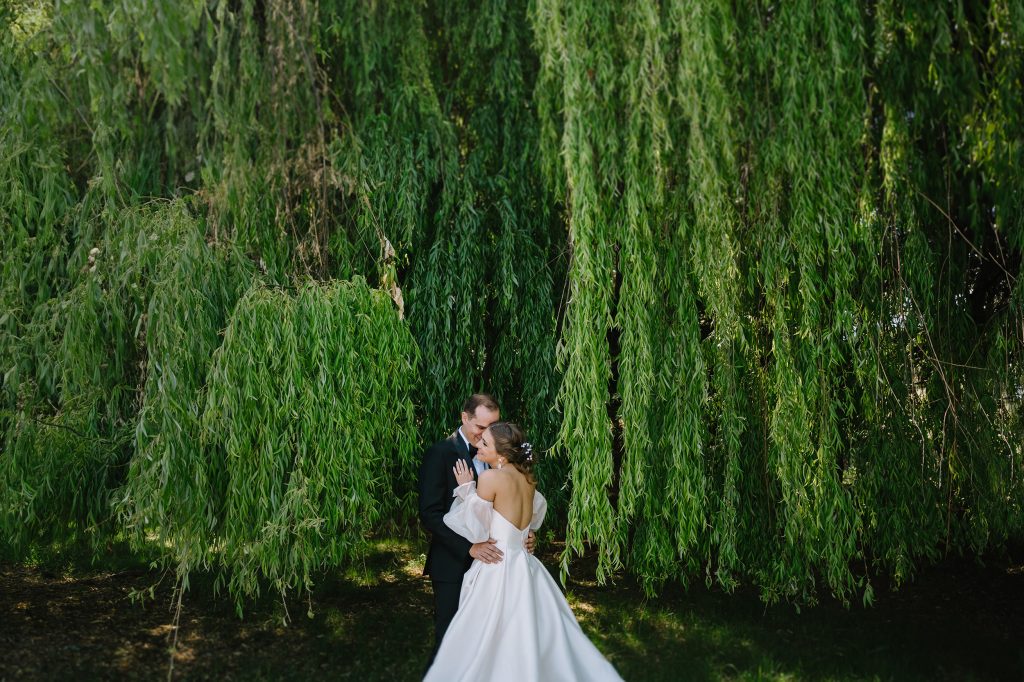 bride and groom embracing on their wedding day by a willow tree at St Mary’s College